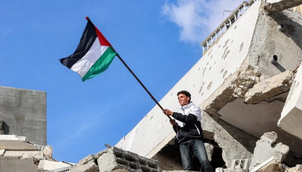 A Palestinian holds the national flag atop the rubble of a collapsed building in the northern Gaza on 19 January, 2025 after a ceasefire deal in the war between Israel and Hamas-afp.jpg